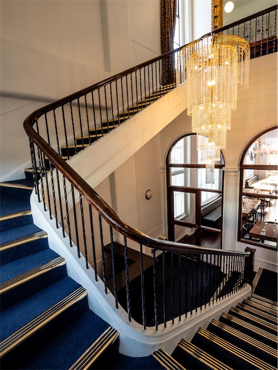 Blue and gold stairs come up toward the camera and turn to the left and then continue up away. They have a dark wood handrail and twisted wrought iron posts.